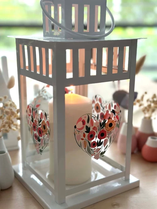 Laterne Hochzeit "Aquarell" aus weiß lackiertem Metall, mit Glaseinsätzen an vier Seiten. Das Bild zeigt die Laterne von schräg hinten. Die Gläser auf der Rückseite und die Seitengläser sind zusehen mit einem Aquarellherz aus Blumen.