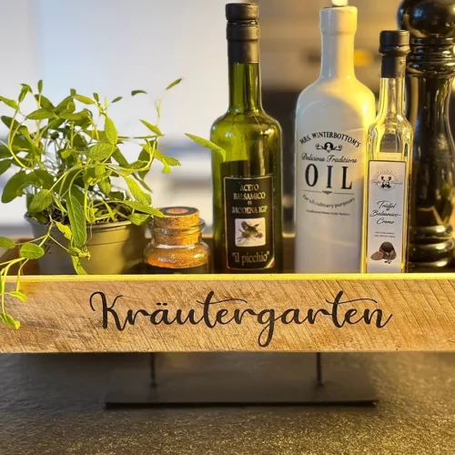 tischboard-dekoboard aus Holz im Shabbystyle, personalisiert mit Schriftzug "Kräutergarten", auf schwarzem Metallsockel, dekoriert auf einer Küchenarbeitsplatte. Im Tischboard dekoriert sind eine Pfeffermuehle, ein Salzstreuer, eine Ölflasche und eine Kraeuterpflanze.