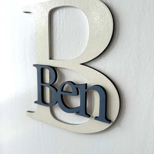 Einzigartige Namensschilder für die Zimmertüre aus MDF, zweifarbig lackiert. Das Türschild besteht aus einem 20 cm großen Buchstaben B und dem ausgeschriebenen Namen Ben, fixiert auf dem Großbuchstaben. Befestigt an einer weißen Türe.