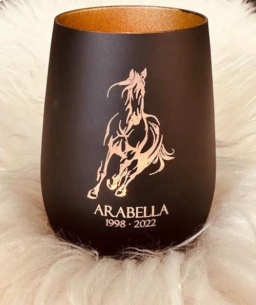 Windlicht Pferd, Glas innen gold, Außen schwarz, mit graviertem Pferdemotiv, Namen des Pferdes, Geburtsjahr und Sterbejahr, dekoriert auf einem weißen Fell