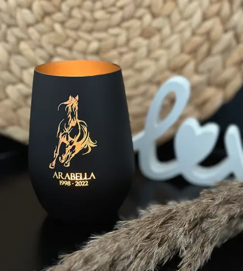 Windlicht Pferd, Glas innen gold, Außen schwarz, mit einem brennenden Teelicht im Glas. Man sieht das Motiv, den Name des Pferdes und das Geburts- und Sterbejahr leuchten.