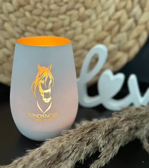 Windlicht Pferd, Glas innen gold, Außen weiß, mit einem brennenden Teelicht im Glas. Man sieht das Motiv, den Name des Pferdes und das Geburts- und Sterbejahr leuchten.